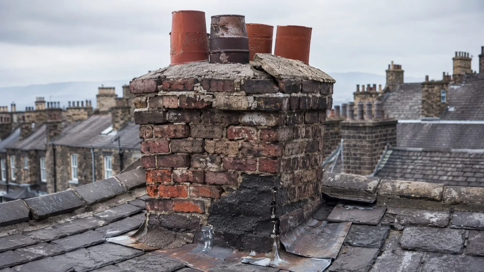 A failing brick chimney stack before work: crumbling mortar joints, spalled bricks, the stack leaning out of plumb and lifting lead flashing at the base.