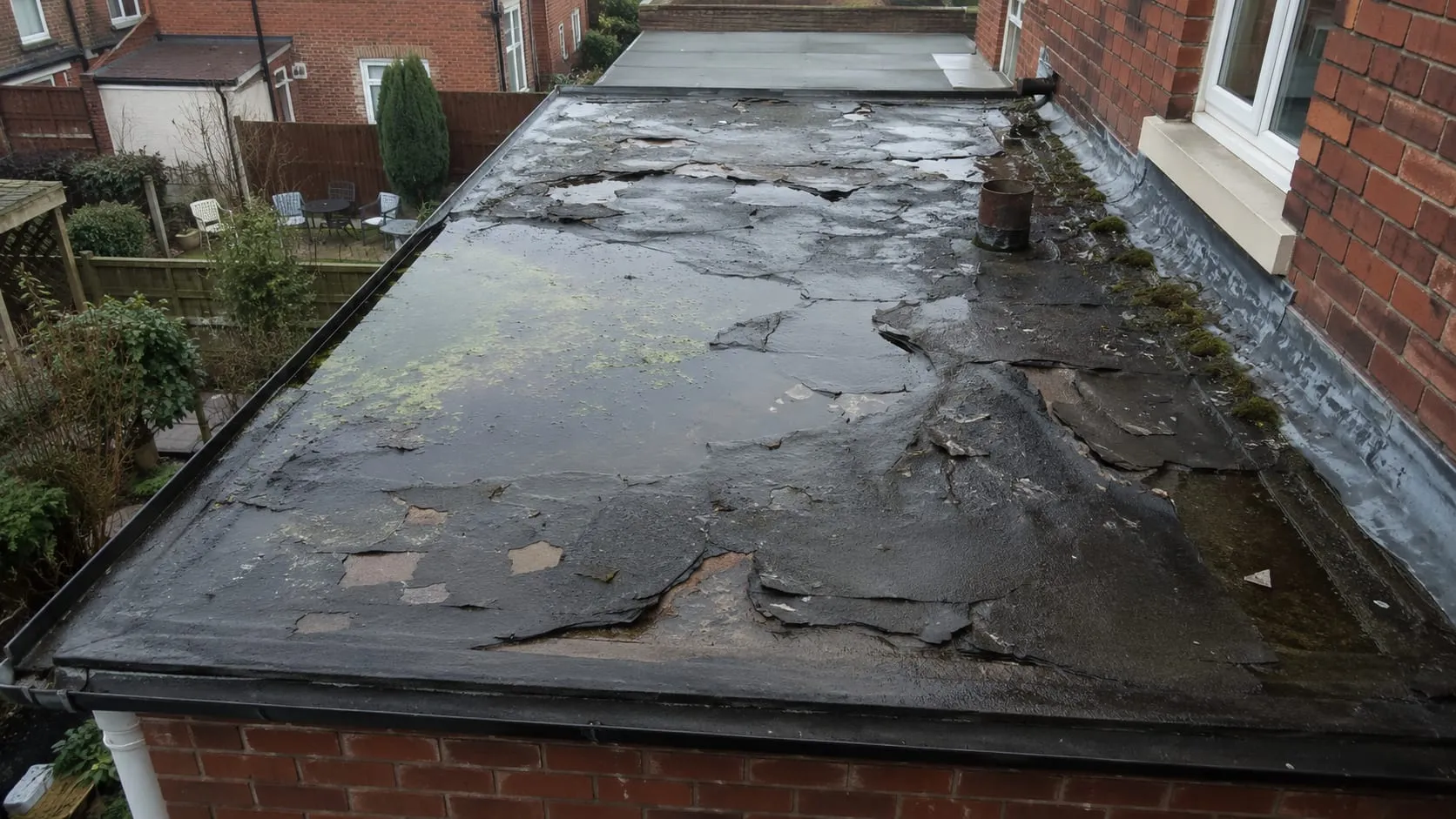 A failing felt flat roof on a single-storey rear extension: blistered and split felt, bitumen patch repairs and rainwater ponding in a low area.