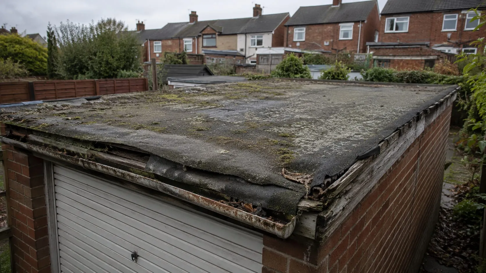 A failing felt garage roof: cracked and shrunken felt splitting at the edges, heavy moss and a rotting timber fascia with a half-detached gutter.
