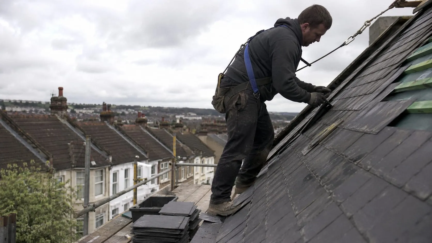 A Ridgeline roofer, harnessed, laying natural slate on a Sheffield rooftop with terraced streets and hills behind